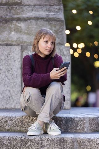 Contemplating on urban stone steps holding phone, young woman in burgundy sweater at dusk