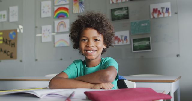 Young African American boy smiling while seated at classroom desk, colorful artwork on walls reflecting creativity and learning. Ideal for educational materials, school promotions, childcare advertisements, or any use representing school environments, academic achievements, and children's educational activities.