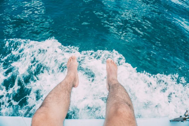 Man swinging bare feet over edge of yacht above clear ocean water, capturing a sense of relaxation and freedom. Ideal for travel blogs, summer vacation advertisements, marine-themed designs, and lifestyle content.