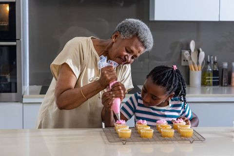 African grandmother guiding granddaughter piping pink frosting on cupcakes in kitchen