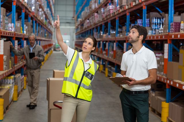 Warehouse workers checking inventory on shelves. Female worker in safety vest pointing while male colleague holds clipboard. Ideal for articles on logistics, supply chain management, warehouse operations, teamwork, and industrial organization.