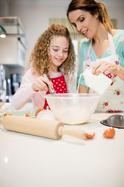 Mother and daughter are standing in a bright kitchen, with the mother pouring milk into a bowl of flour while the daughter uses a whisk. They are both wearing aprons and smiling, enjoying the cooking process. Kitchen utensils and ingredients are visible on the countertop, creating a homely and engaging atmosphere. This stock photo is ideal for content related to family activities, cooking tutorials, parenting blogs, and advertisements showing home life and family bonding.