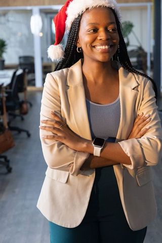 African american businesswoman standing, smiling in santa hat with arms crossed in office