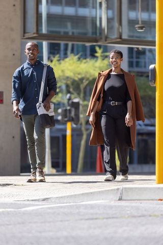African american couple walking sunlit city crosswalk in brown coat with messenger bag