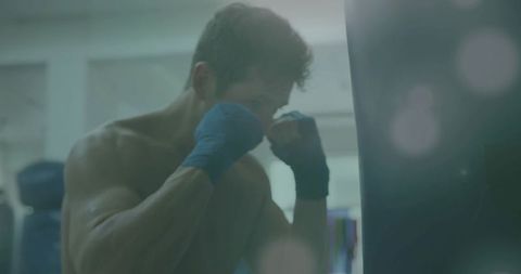 Shirtless boxer training on heavy bag with blue hand wraps showing focus and power