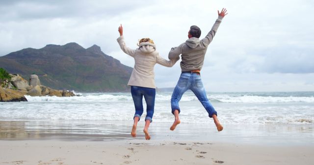 Couple jumping mid-air on sandy beach with mountains and ocean in background. Both wear casual clothing. Use for themes related to travel, vacations, relationships, outdoor activities, happiness.