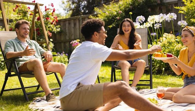 Friends sharing fruit and laughing at sunlit backyard picnic on blanket and lawn