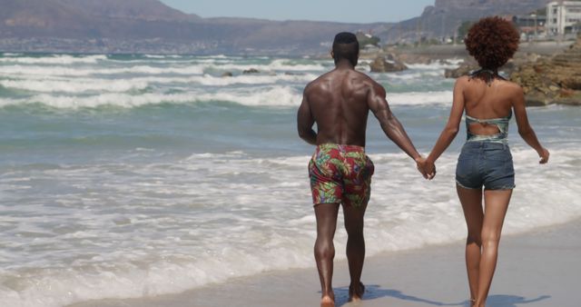 Diverse couple walking and holding hands by sea on sunny beach. Togetherness, summer, vacations and free time, unaltered.