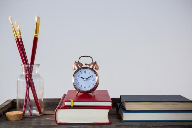 Books, an alarm clock, and paint brushes are neatly arranged on a wooden table. This image can be used to represent themes of education, creativity, and time management. Ideal for use in educational materials, art-related content, or organizational tips.