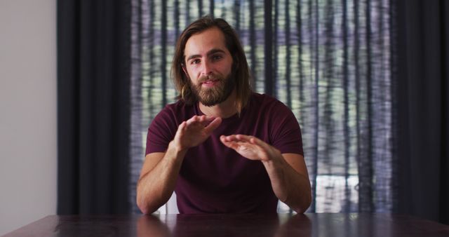 Young man with long hair and beard sits at a table indoors, gesturing with his hands as if discussing or explaining a concept. The casual setting with curtains in the background makes it suitable for use in content about online discussions, educational presentations, or casual conversation topics.