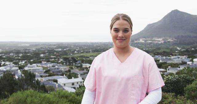 Young nurse wearing pink uniform standing outdoors with a scenic view of a town and mountains in the background. Ideal for use in healthcare articles, promotional material for medical services, outdoor relaxation, or community health initiatives.