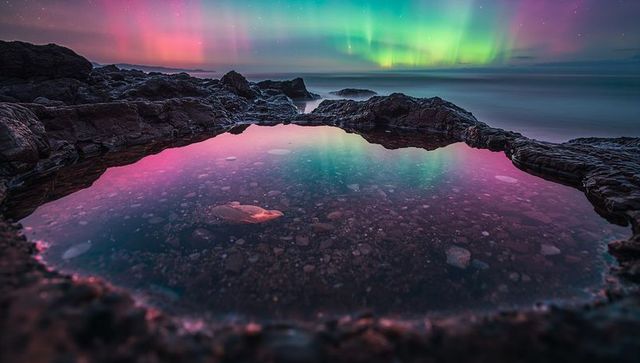 Reflecting tide pool mirroring vibrant aurora borealis over rocky coastal nightscape