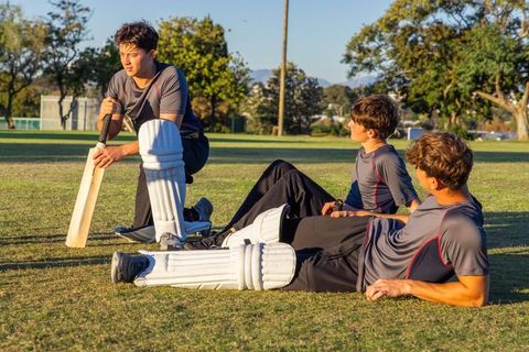 Teen male cricket players resting on grass field during golden hour training break
