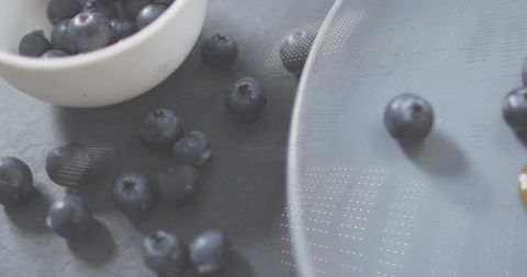 Scattering blueberries on dark slate with white bowl and matte grey ceramic plate