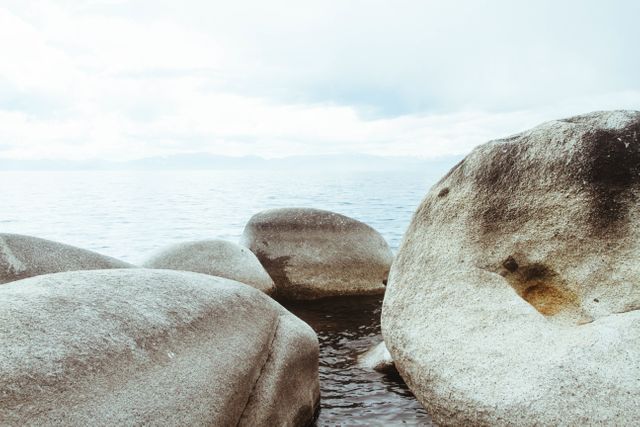 Calm and serene scene capturing large rocks on a shoreline overlooking tranquil waters under a partly cloudy sky. Ideal for projects requiring peaceful and natural landscapes. Suitable for nature-themed prints, travel promotions, or relaxation content.