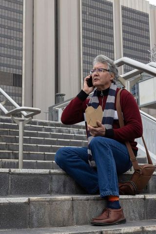 Senior man eating takeout on city stairs talking on phone during urban lunch break with scarf