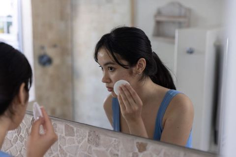 Young asian woman pressing cotton pad to cheek at bathroom mirror for skincare routine