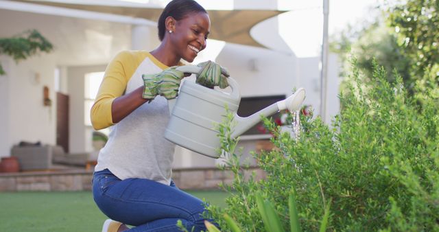 Perfect for articles and promotional materials about gardening, outdoor hobbies, sustainable living, or home improvement. This image of a smiling woman watering plants wearing gloves and holding a watering can captures a joyful moment in a sunny backyard garden.