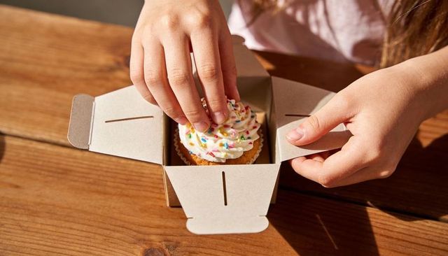 Child Lowering Frosted Cupcake with Sprinkles into Cardboard Bakery Box on Wooden Table