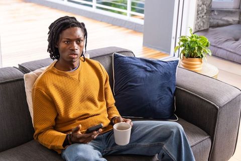 African american man sitting on sofa holding smartphone and mug in modern living room