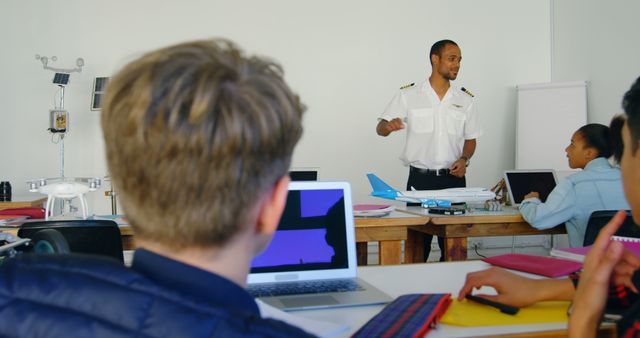 Aviation instructor leading a classroom session with students. The instructor is in uniform, discussing topics in aviation. Students are using laptops and models of aircraft for hands-on learning. This image can be used for educational materials, aviation training programs, coaching sessions, academic articles on aviation education, and more.