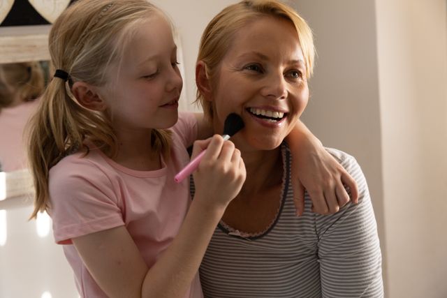 Caucasian woman spending family time together with her daughter at home, the girl holding makeup brush doing her mothers makeup. Family leisure time at home.