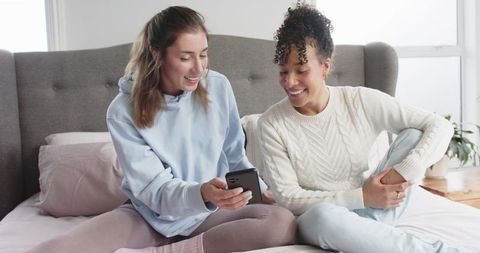 Two Friends Sharing Smartphone on Cozy Bed Smiling and Laughing Casual Home Lifestyle