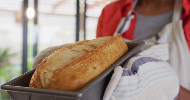 Close-up of person holding a freshly baked artisan bread in loaf pan with oven mitt in kitchen. Ideal for promoting home cooking, baking recipes, culinary blogs, and cookbooks. The warm lighting and homey feel enhance themes of freshness and homemade goodness.