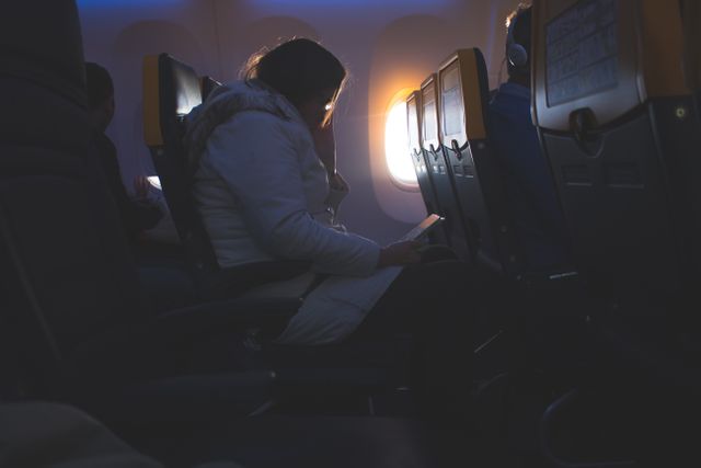 Passenger engrossed in tablet during flight, captured in soft lighting from the airplane window. Ideal for depicting modern air travel, inflight entertainment and use of digital devices while flying.