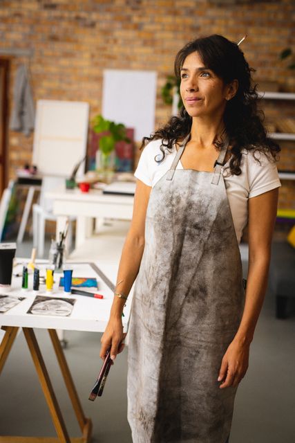 This image shows a smiling biracial female artist standing in her creative studio, wearing an apron and holding painting supplies. Ideal for use in articles or advertisements related to art, creativity, artist profiles, and artistic workshops.