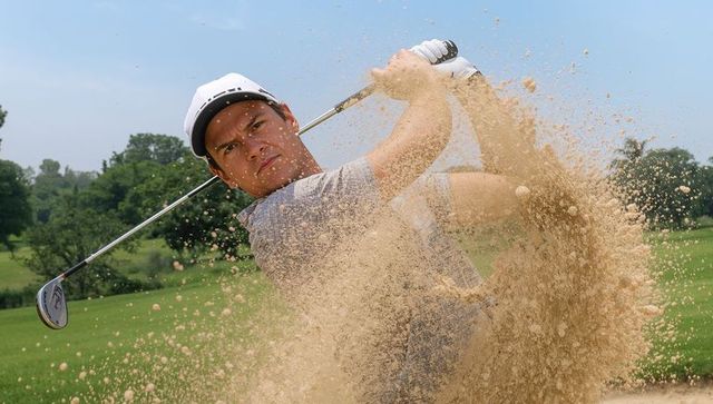 Golfer hitting explosive bunker shot with sand spray, iron club, white cap and glove