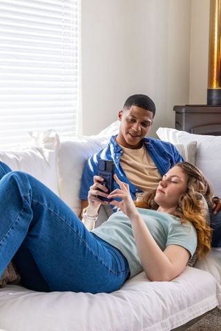 African american man and woman relaxing on white couch sharing smartphone in bright home