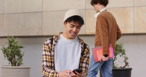 Asian university student smiling and texting on campus plaza with backpack and beanie