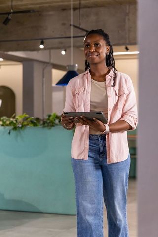 African american woman standing in coworking space holding tablet, wearing pink shirt