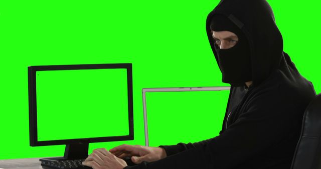 A Caucasian man in a hoodie appears to be engaging in secretive computer work, with copy space on the green screen monitors. His attire and the green screens suggest a theme related to hacking or cybersecurity.