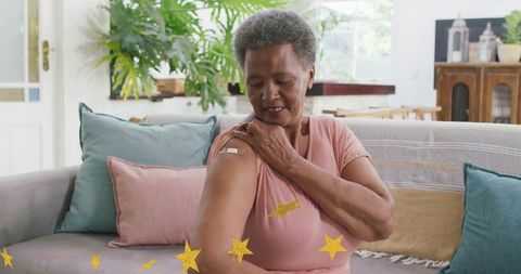 Senior woman touching vaccinated upper arm while sitting on sunlit sofa at home