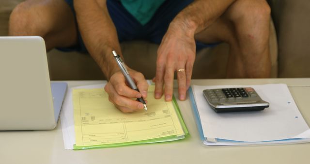 A middle-aged Caucasian man is seen filling out paperwork, related to finances or taxes, with a calculator and laptop nearby, with copy space. His focus on the task suggests a setting of personal accounting or business administration.