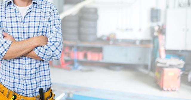 Mechanic confidently standing in a garage with folded arms, wearing tool belt and plaid shirt. Background is blurred, focusing on the professional demeanor. Suitable for promoting automotive services, mechanic training courses, maintenance workshops, and automotive industry advertisements. Ideal for highlighting confidence and reliability in vehicle repair fields.