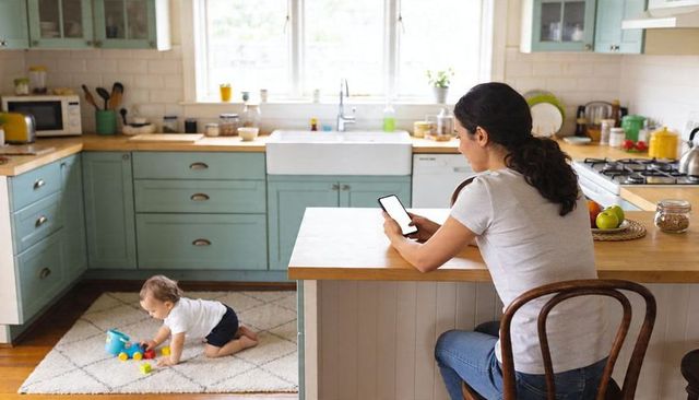 Mom checking smartphone while baby playing on rug in sunlit farmhouse kitchen