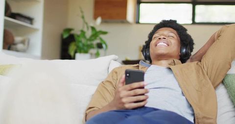 Young man relaxing on modern sofa wearing headphones enjoying music with smartphone