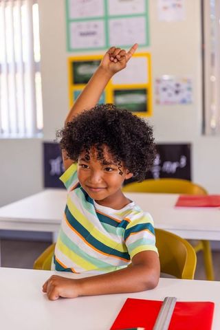 African american boy raising hand in classroom at desk with red folder and ruler