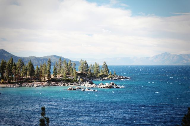 Depicts a panoramic scene of Lake Tahoe with clear blue water, surrounded by a forest of pine trees and distant mountains under a partially cloudy sky. Ideal for travel magazines, nature blogs, tourism brochures, and outdoor recreational advertisements.