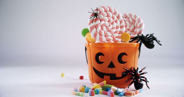 Halloween-themed bucket filled with lollipops and surrounded by colorful candies and artificial black spiders. Ideal for use in festive advertising, holiday promotional material, or as a visually appealing element in Halloween party invitations and decor.