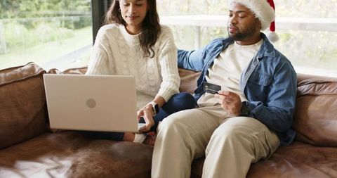 Couple shopping online on sofa with laptop, man wearing santa hat holding credit card