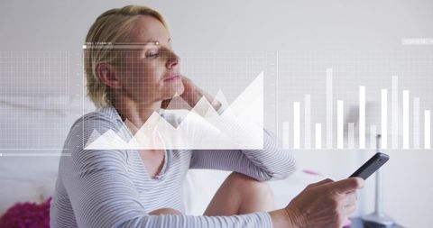 Mature woman holding smartphone and reflecting in bedroom with overlaid analytics graphs