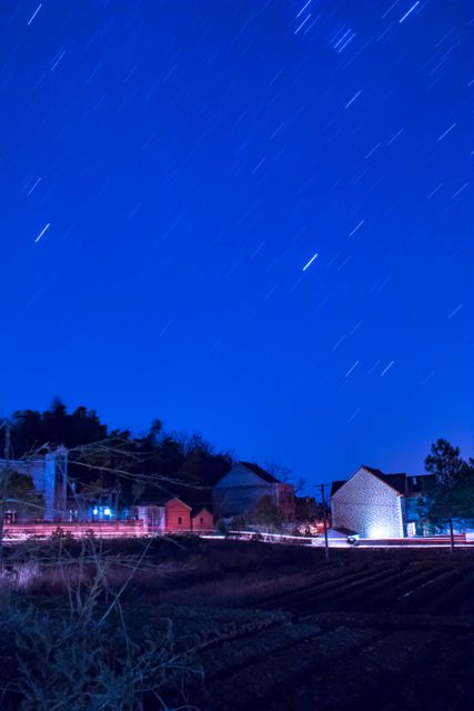 View of rural countryside under night sky with star trails. Lights from village houses and long exposure create an ethereal scene. Ideal for use in articles on rural life, night photography, serenity, calm and agricultural topics. Can be used as background image for social media, blogs and websites focused on nature and night sky watching.