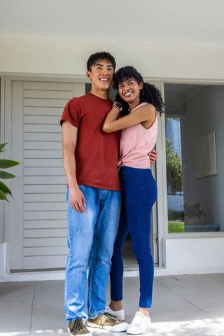 Young diverse couple embracing on porch and smiling while showing smartwatch near home