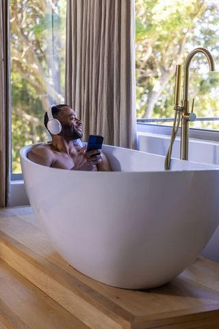 Mature man relaxing in freestanding bathtub with headphones and smartphone, sunlit retreat