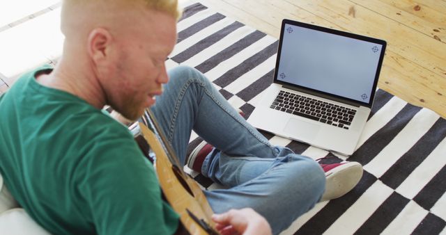 Man practicing guitar at home while using a laptop for online lessons. Perfect for concepts on online learning, home-based hobbies, and blending traditional skills with modern technology.