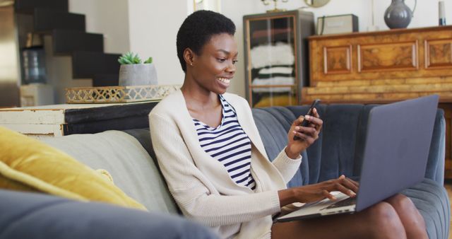 Happy african american woman sitting on sofa in living room, using laptop and smartphone. domestic life, spending time at home.
